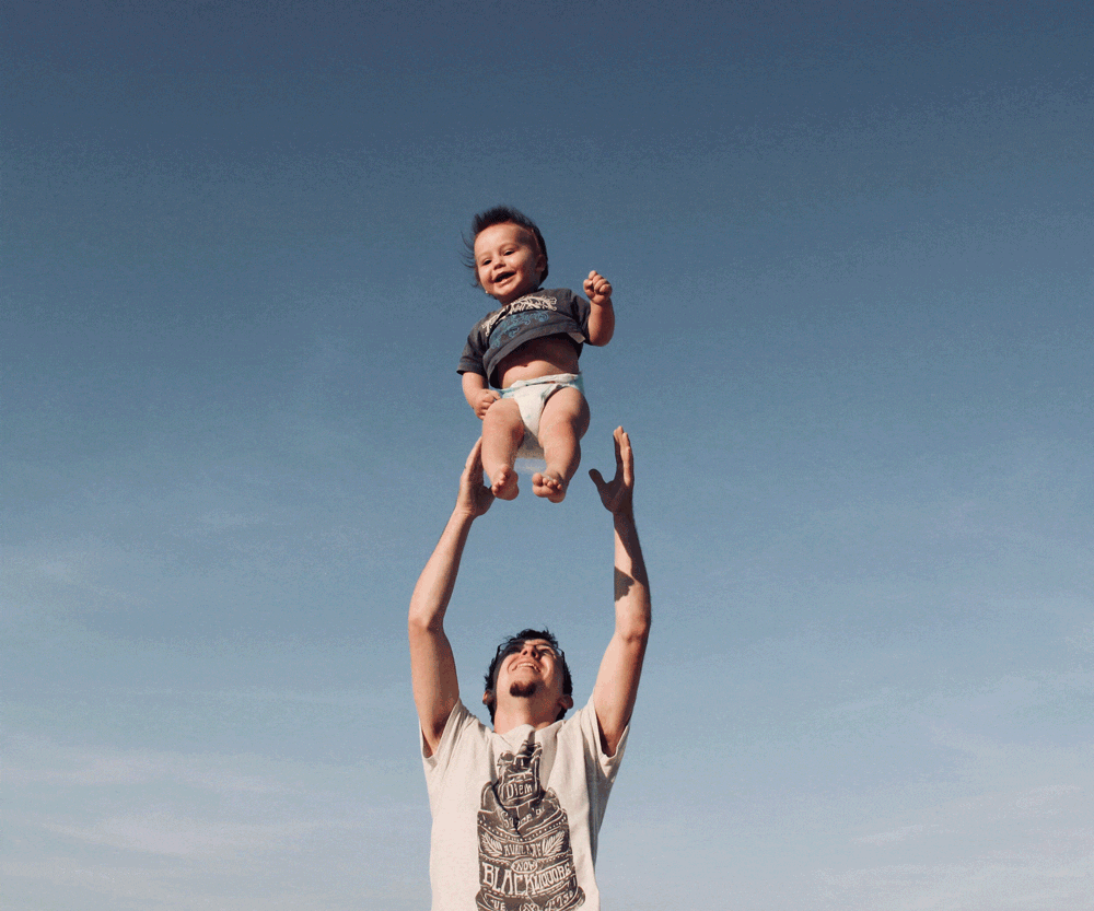 A dad throws a small toddler son outfitted in a t-shirt and diaper into the air outdoors. 