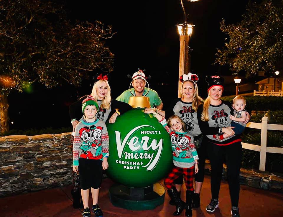 A multigenerational family of seven stands smiling around a huge green Christmas bulb reading "Mickey's Very Merry Christmas Party".