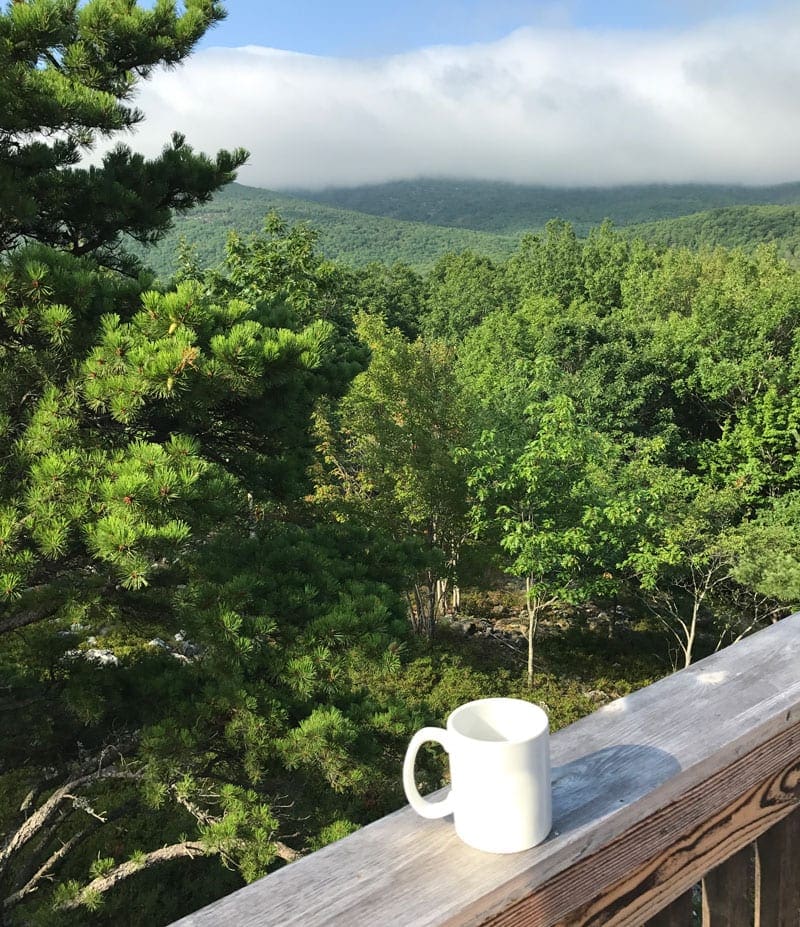 A white coffee mug rests on a deck railing with an exapansive view of the Acadia National Park in the background.