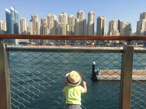 A young boy wearing a yellow shirt and hat looks on at the Dubai skyline.