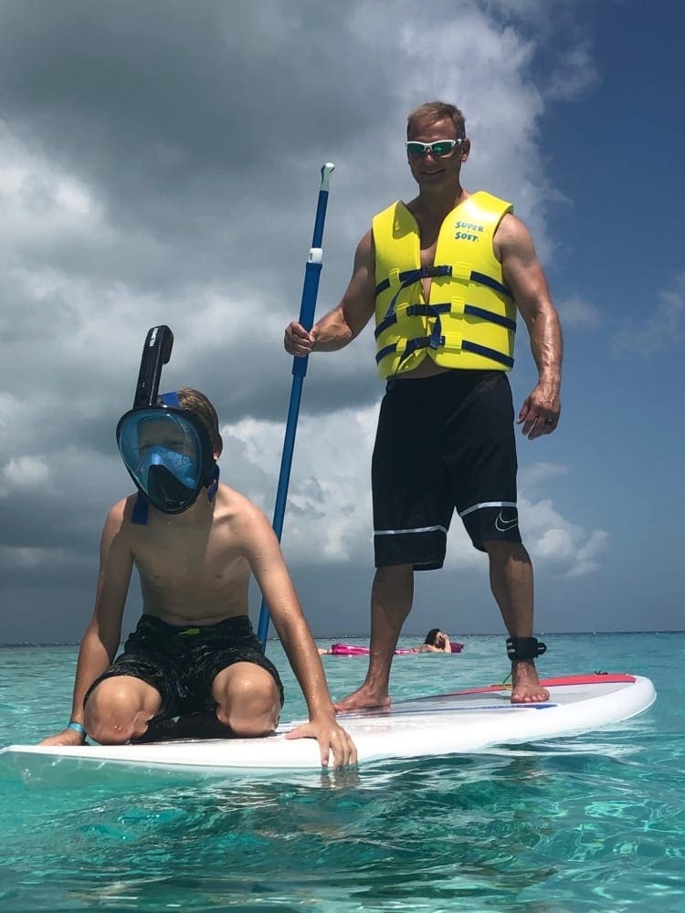 A father stands on a paddle-board, while his young son wearing a snorkel mask sits in front of him, while float along on the waves off-shore from Turks and Caicos.