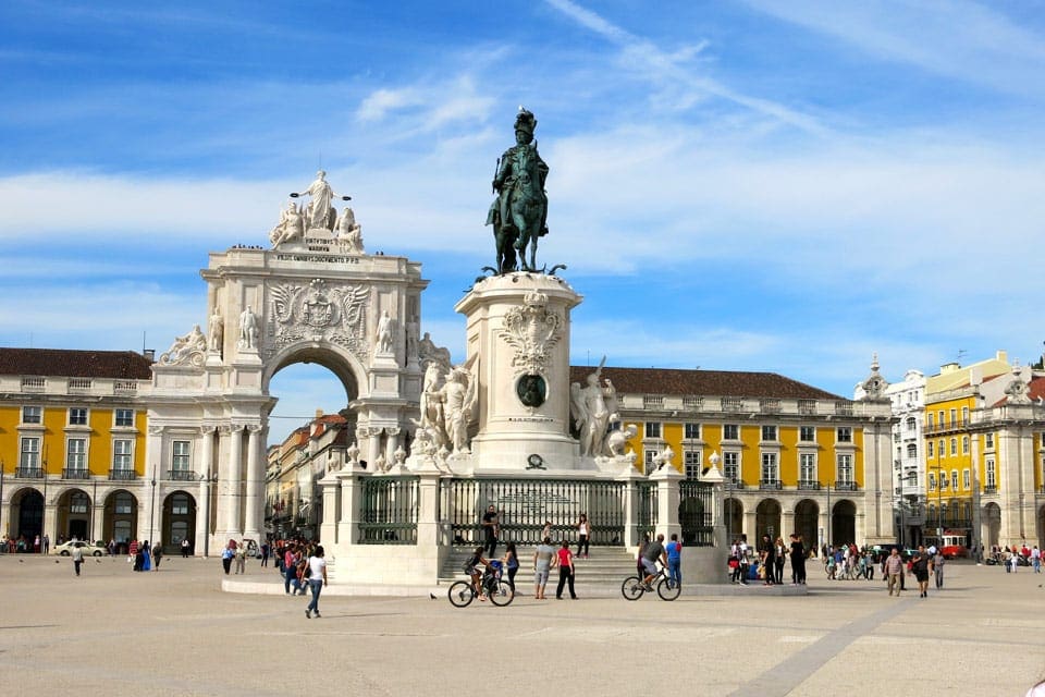 Several people wander amongst the statues and yellow buildings within Praca do Comercio in Portugal.