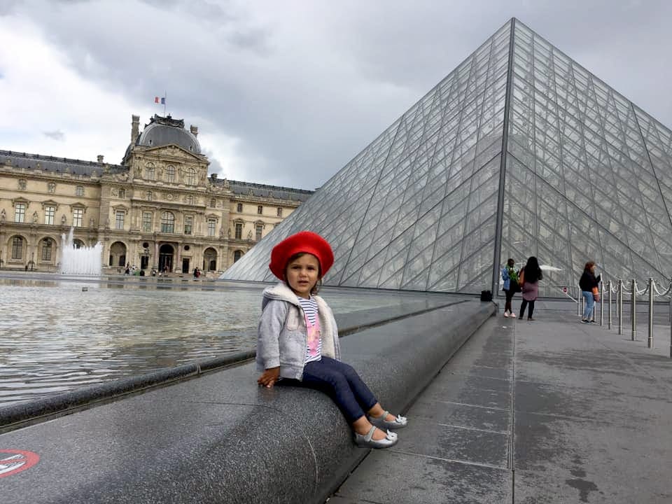 Young girl sits in front of the Louve Museum, one of the stops on our virtual vacation from home to paris