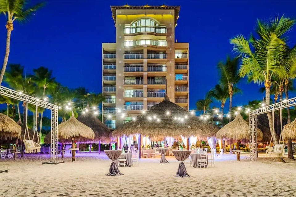 A night view of Hyatt Regency Aruba Resort, Spa and Casino, one of the best family resorts in Aruba, featuring palm trees, sand, and a well-lit cabana.