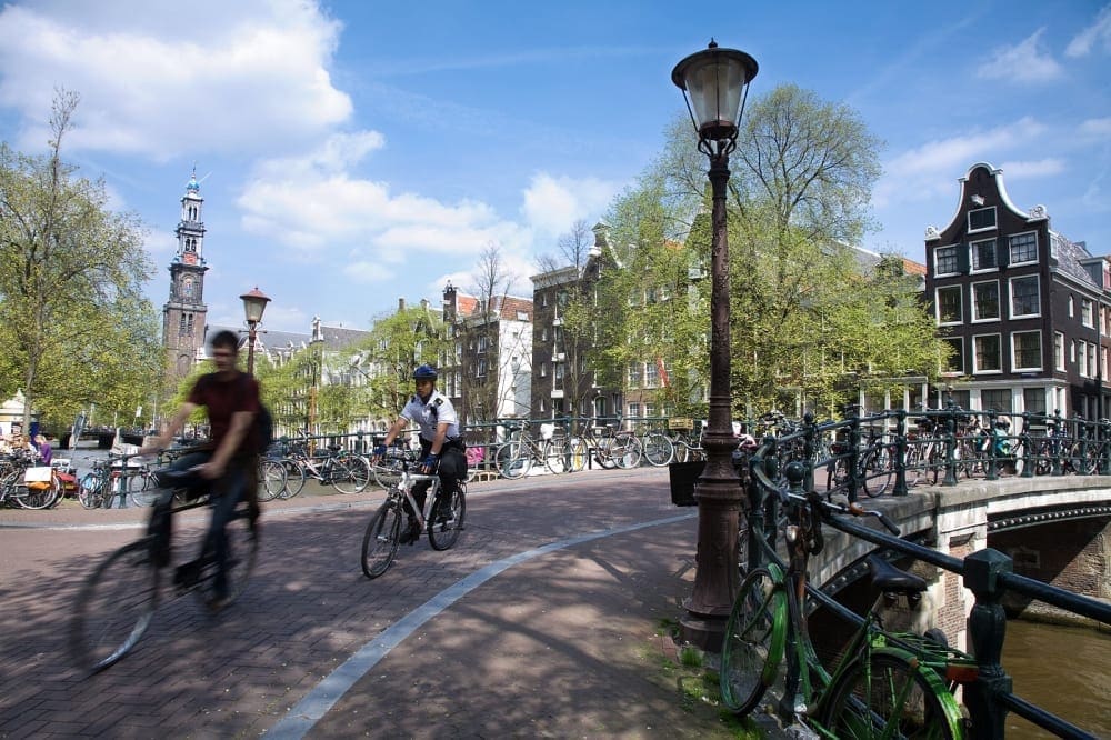 Bikers whizzing by a park in Amsterdam. Biking is a great way to get active on our Virtual Vacation from Home to Amsterdam.
