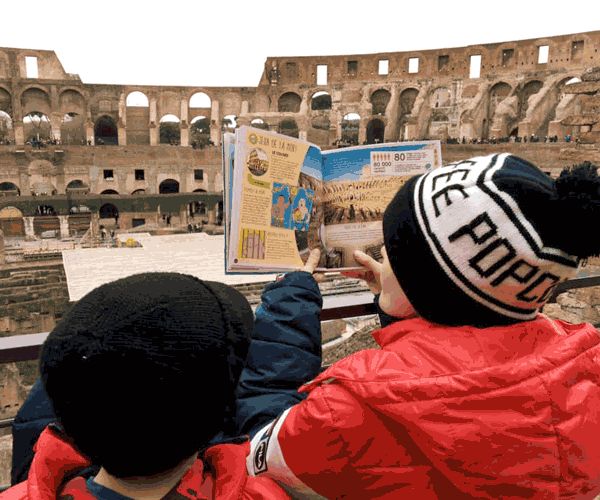 Two boys in looking at a travel book on the Colosseum, while standing inside the Colosseum.