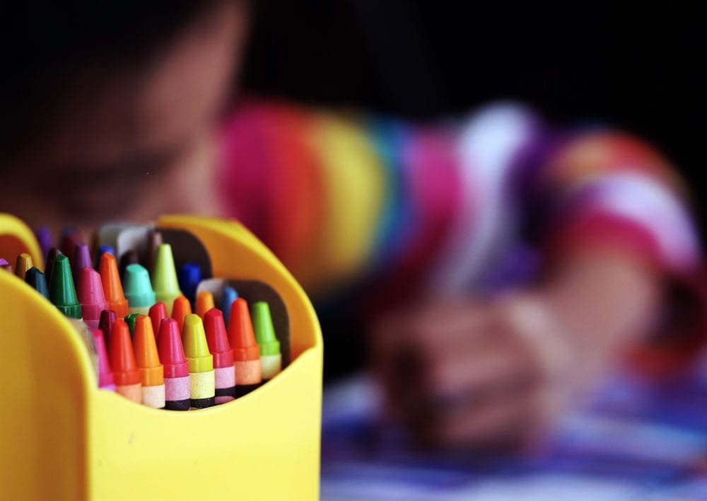A large box of crayon stands in the foreground, with a young child coloring in the background. An art project is a great way to take a virtual vacation to Lucerne.