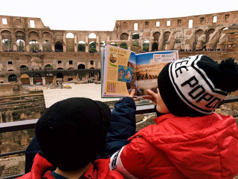 Two boys in looking at a book of the Colosseum, while standing inside of it, a must stop on our Rome itinerary with kids.