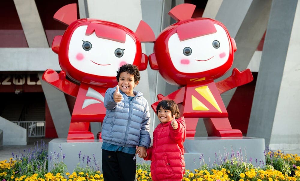 Two boys of color stand in front of two red robots in China. Both boys are smiling at the camera and giving a thumbs up.
