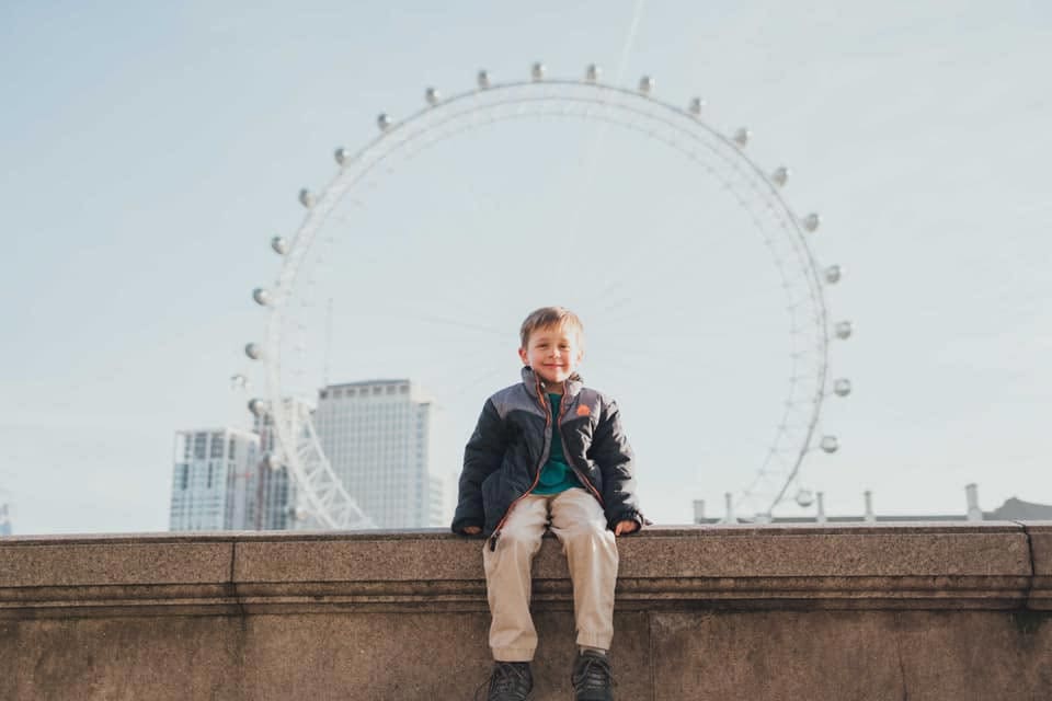 A young boy sits with the London Eye in the distance.