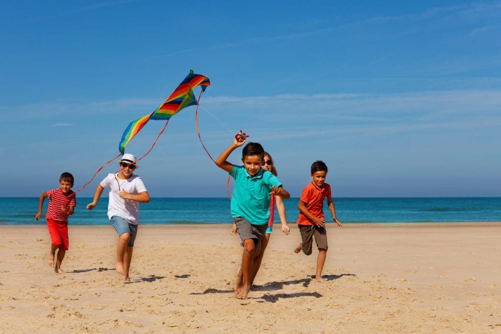 Group of happy children boys and girls run on the sand beach holding colorful flying kite with sea on background.