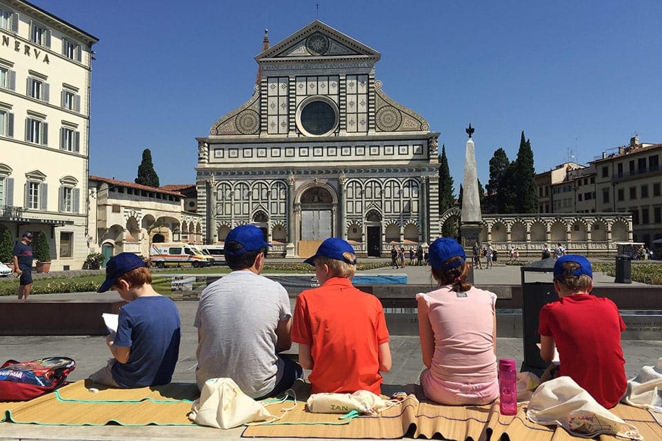 Five kids sitting together eating lunch with a view of an old cathedral with Arte al Sole.