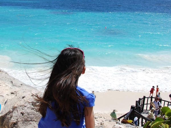 A woman with long dark hair stares out onto the bright blue ocean of Tulum.