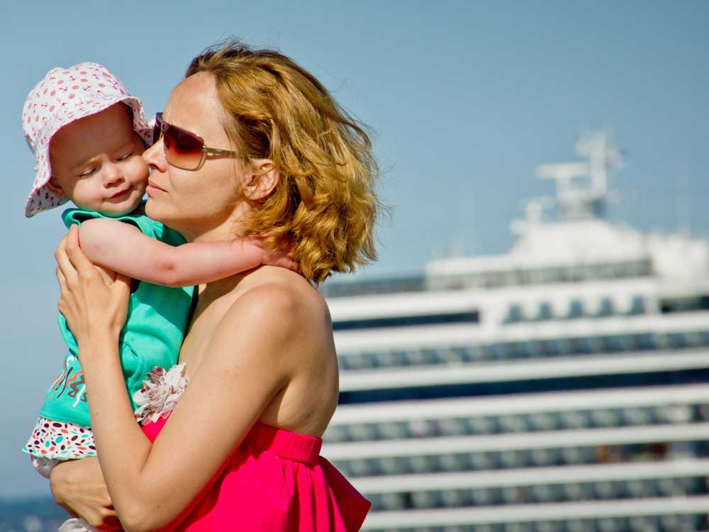A monther hugs her young infant, with a large cruise ship in the background.