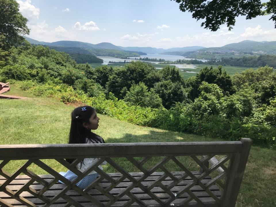 A young girl sitting on the bench facing the lake and the hills of Cold Spring.