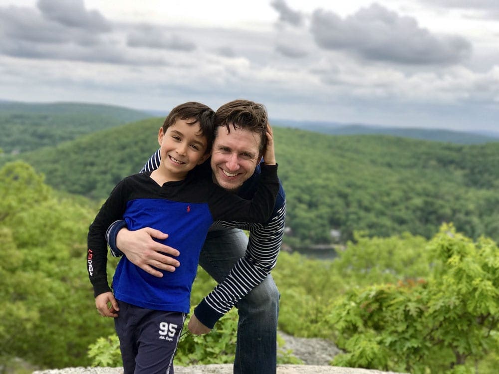 Father and son pose together on top of a mountainside in Litchfield, Connecticut, with an expansive view of lush greens behind them.