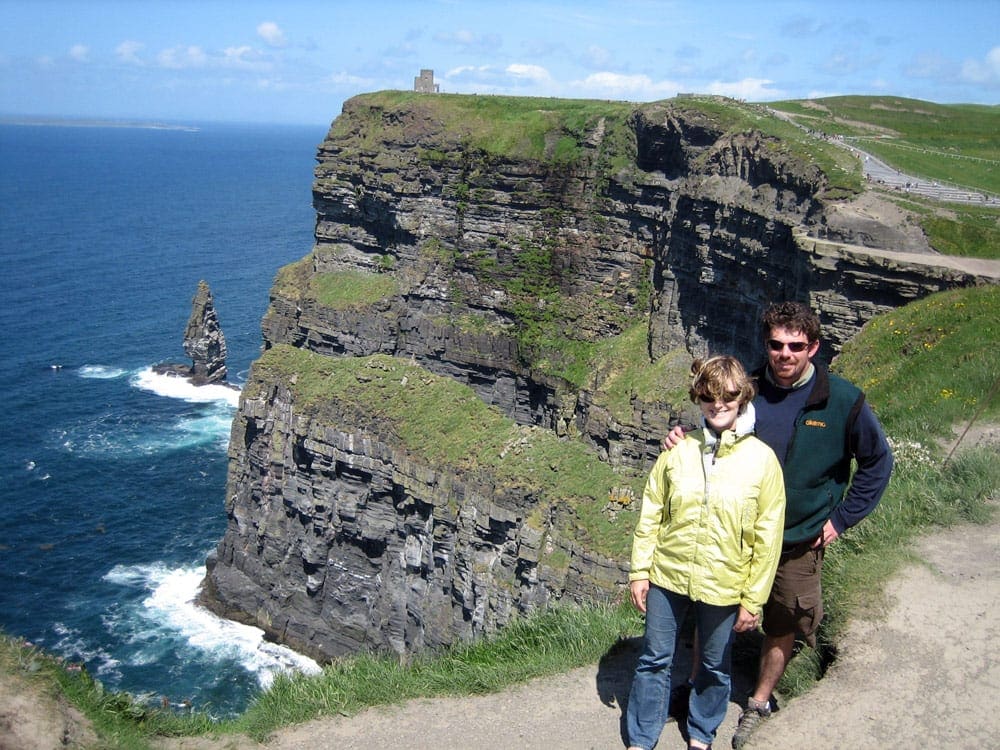 Two adults stand along a cliff in Ireland. The Cliffs of Moher is one of our stops on the virtual vacation to Ireland.