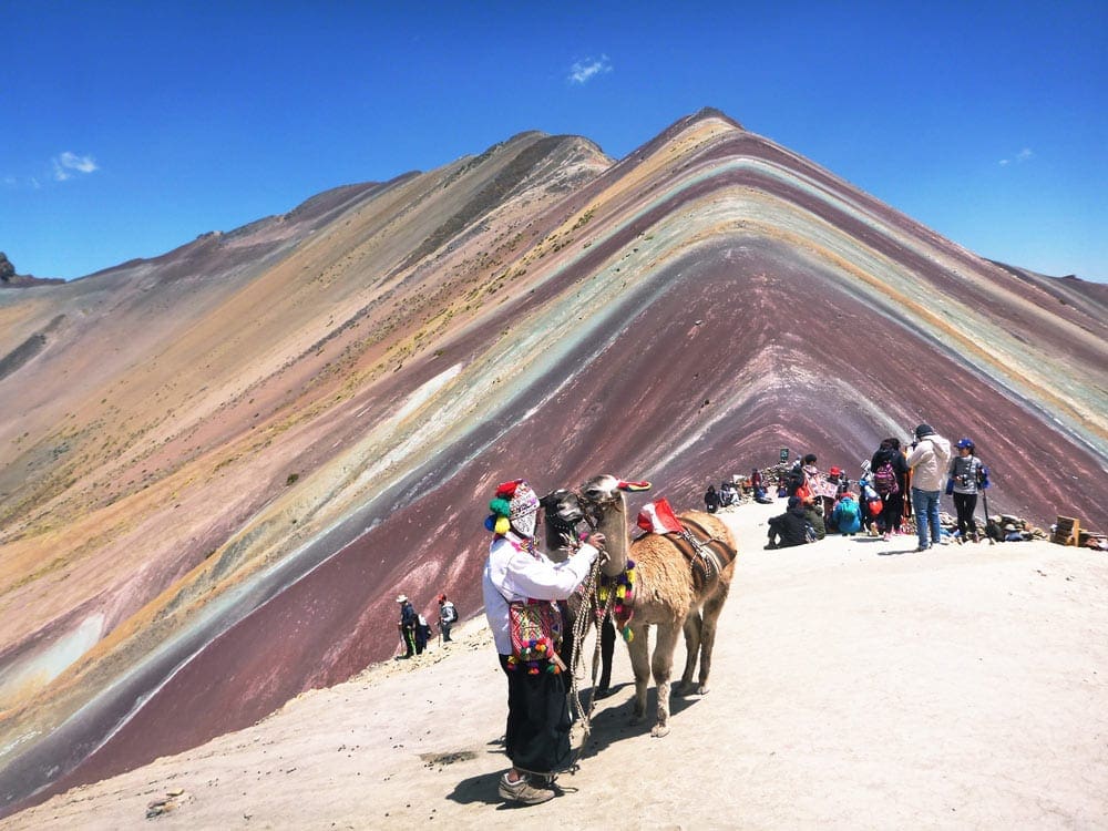 A tour of people walk across the dunes, with Rainbow Mountain in the distance, a great stop on our Peru family vacation itinerary.