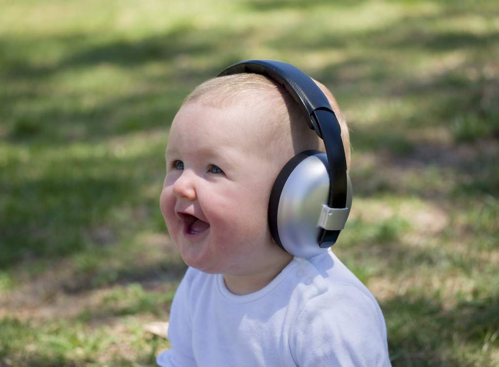 A baby boy wears noise canceling headphones on a sunny day in the grass.