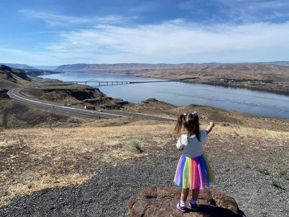 Young girl in rainbow skirt points to river along a road. Taking breaks is a great way for staying sane on family road trips.