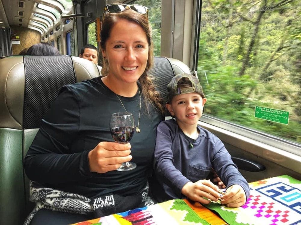 Mother and son sit smiling on a train within the Sacred Valley area of Peru.  Mom is holding a glass of wine. Treating yourself is one of the best tips for traveling with a toddler.