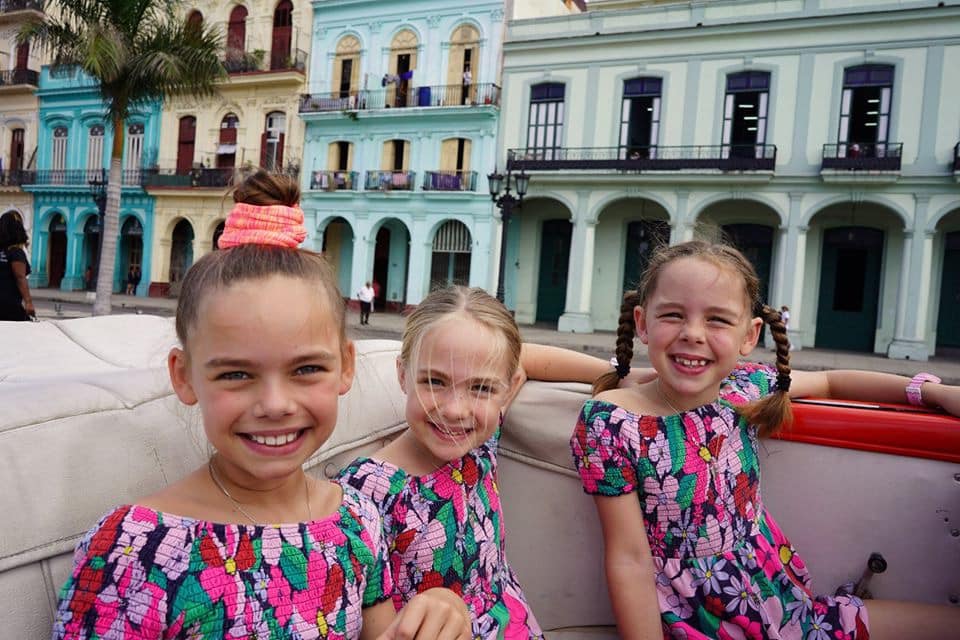 Three young girls, all wearing brighly patterned dresses ride in a corvet along a street in Havana, Cuba.
