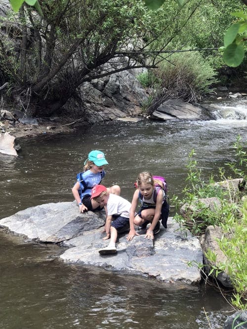 Three young children sit on a large rock in the middle of a river while hiking near Denver.