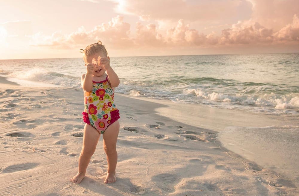Toddler girl in red floral bathing suit playing on sand at Navarre Beach in Florida, one of the top places in travel in 2023 with kids.