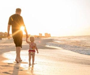 A father holds the hand of his toddler while walking along Navarre Beach at sunset.