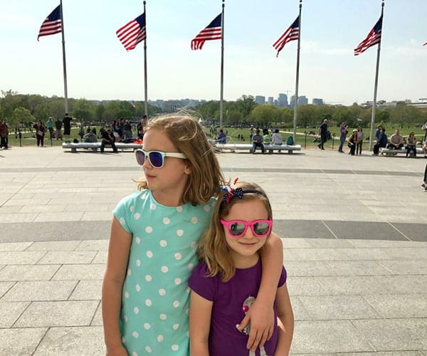 Two girls stand smithing with sunglasses on in front of five American flags in Washington DC, one of the best Fourth of July destinations for a family trip.