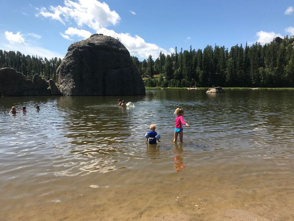 Two young kids play in a lake with a large bolder and row of evergreen trees in the background on the grounds of Custer State Park in South Dakota, one of the best affordable summer vacations in the United States with kids.