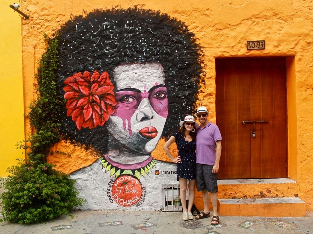 A young couple stands smiling next to an expansive piece of street art in Cartagena, Colombia, featuring a large women's head with an afro and a red flower behind her ear.