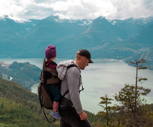 A man hikes along a trail with a young girl on his back in a carrier, with a large lake and mountains in the background.