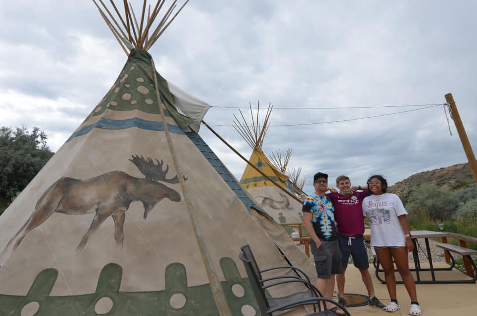 Three people stand smiling to the right of a large teepee for glamping.