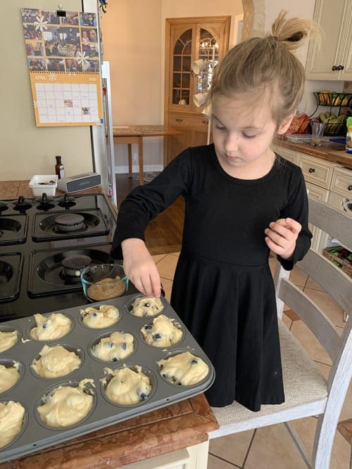 Young girl putting sugar on top of blueberry muffins.