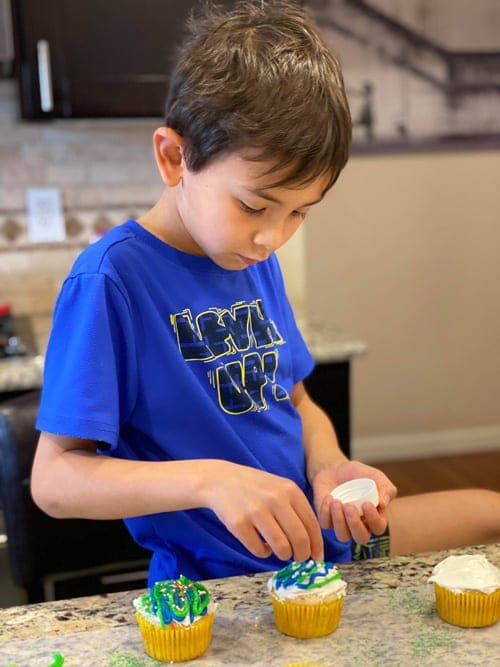 Young boy decorates a vanilla cupcake with blue and green sprinkles.