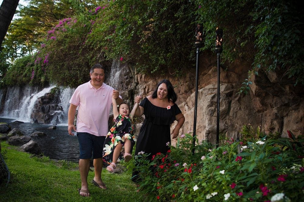 Parents swing their young daughter while walking in a lush Hawaiian park filled with flowers and a waterfall in the backfound.