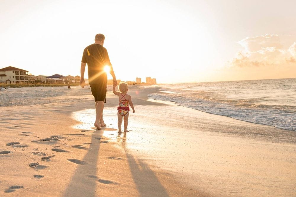 A father holds the hand of his toddler while walking along Navarre Beach at sunset.