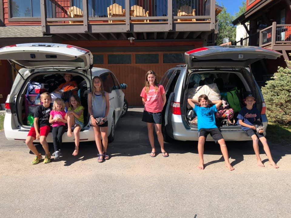 7 kids stand in front of 2 packed vans in preparation of a road trip.
