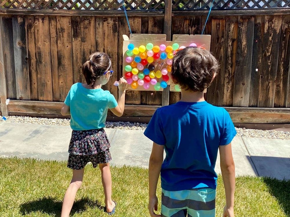 Two young kids use darts to pop a display of balloons in a backyard carnival.