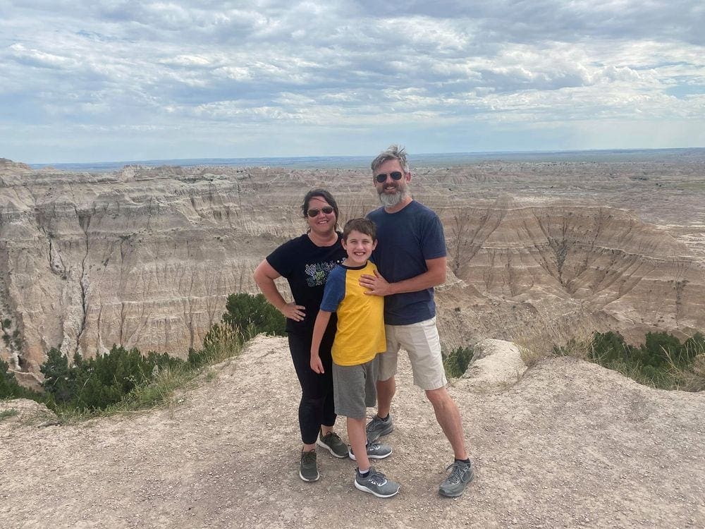 Family of three stands with an expansive view of the Badlands in the background in South Dakota, one of the best affordable summer vacations in the United States with kids.