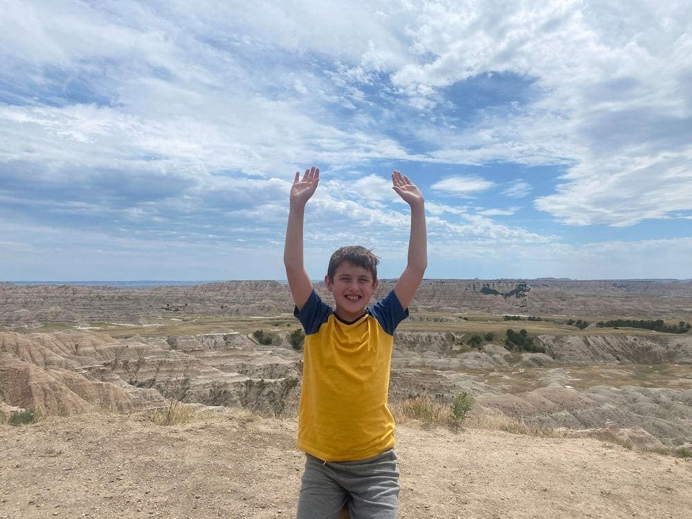 A 4th grade boy stands excitedly with hands in the air in the Badlands.