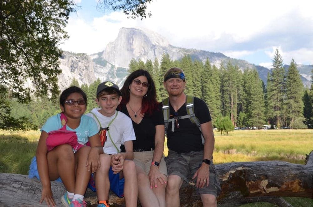 A family of four sits upon a log in front of a mountain view at Yosemite National Park, one of the best west coast national parks for kids!