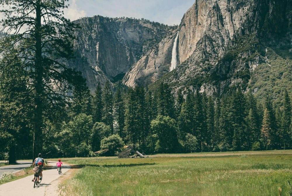 A dad and two children bike on a path within Yosemite Natioanl Park, large trees and mountains dot the background.