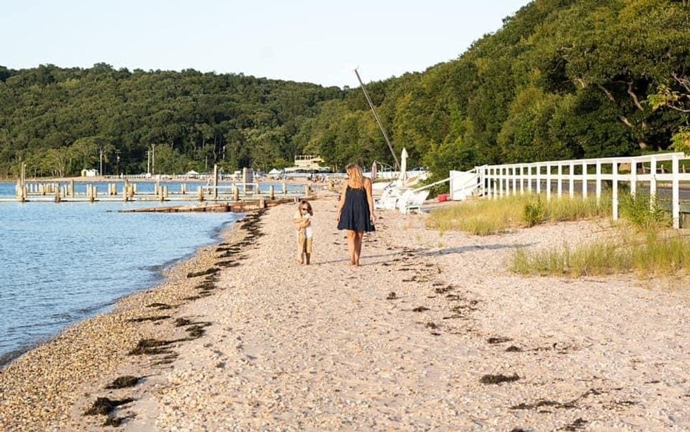 A mom wearing a black dress looks on at her young daughter in white as they walk down a beach. There is water on the left and grassy patches with trees on the right.