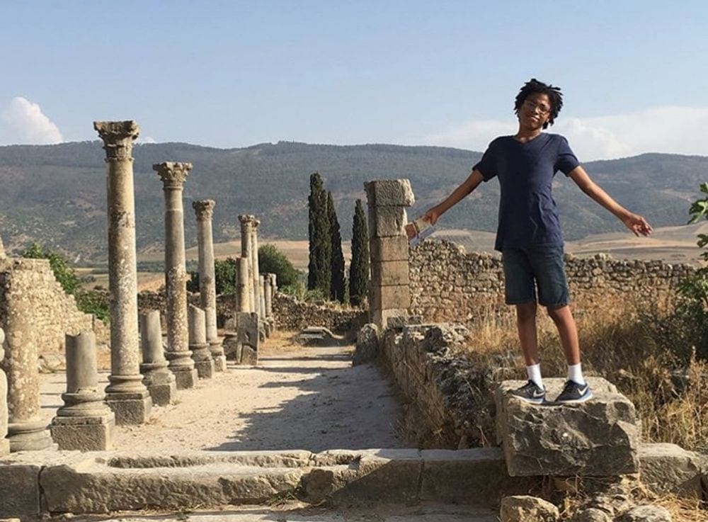 A teenage black boy stands on a large stone at the Volubilis Roman ruins in Northern African. Several ancient columns and stones walls dot the background. Learning around the world is one of the benefits of traveling with kids.