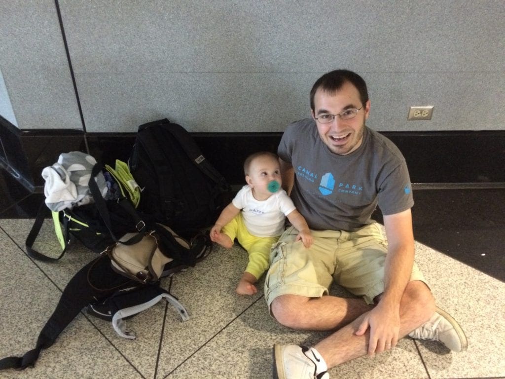 An infant daughter looks up at her smiling dad while sitting on the floor of an airport. Their luggage sits nearby in a big heap.
