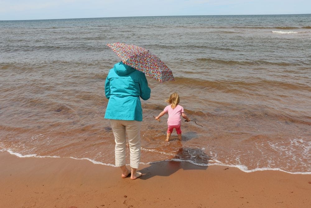 A great-grandmother stands in splashing waves as her granddaughter splashes around on a beach in Prince Edward Island.
