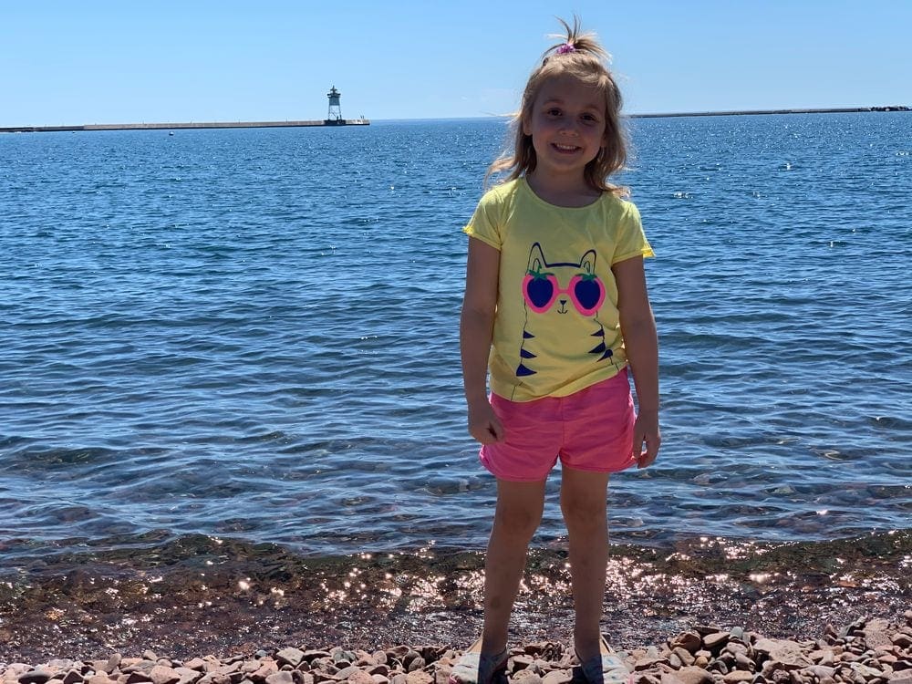 A young girl wearing a yellow shirt stands on a pebbled beach in front of Lake Superior with a lighthouse in the background. Exploring your local area is a great way to recharge your batteries as a family.