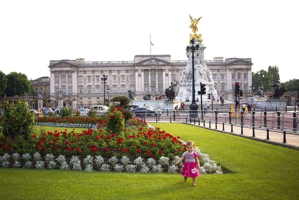 An infant girl takes baby steps in front of a stunning garden outside of Buckinham Palace, shown in full in the background.